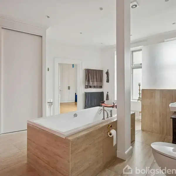 Badekar står i et lyst badeværelse med hvide vægge og trægulv. Håndvask og toilet er placeret mod højre, og håndklæder hænger ved siden af en bruser i baggrunden.