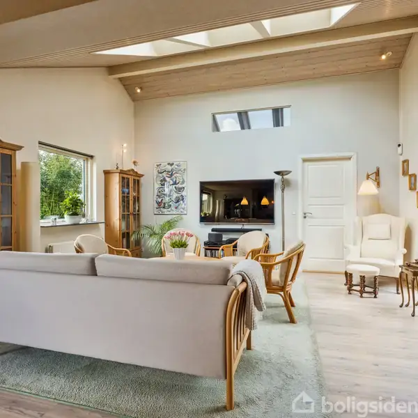 Stue med en hvid sofa og træmøbler, arrangeret omkring et sofabord. Et stort vindue og ovenlysvinduer lader lys ind i rummet, som har en lys og indbydende atmosfære.