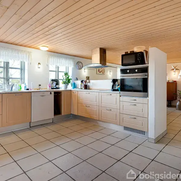 Køkken med lyse træskabe og hvidevarer, herunder ovn og opvaskemaskine. Flere planter placeret på bordpladen. Træloft og flisegulv. Dør fører til anden del af huset.