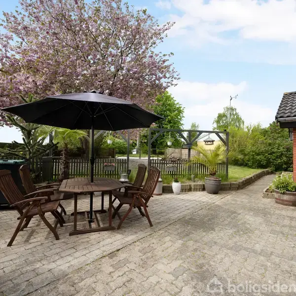 Et bord med stole står under en sort parasol på en terrasse med brosten. Omkring er der blomstrende træer, planter i krukker og udsigt til en grøn have foran et murstenshus.