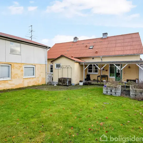 Et gult hus med rødt tegltag står stille i en have med grøn græsplæne og stensætning. Til venstre ses et tilstødende bygning. Vejret er klart med blå himmel.