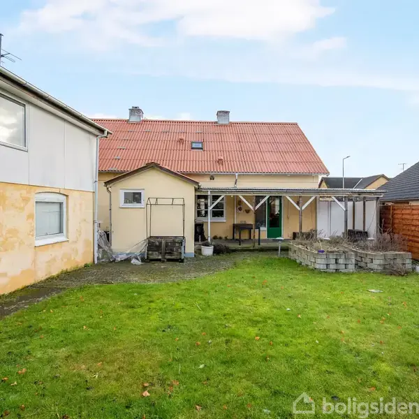Hus med rødt tag og flere skorstene, stående stille i en grøn have omkranset af hegn og bygninger. Terrasse med overdækning foran huset, omgivet af en velholdt græsplæne.