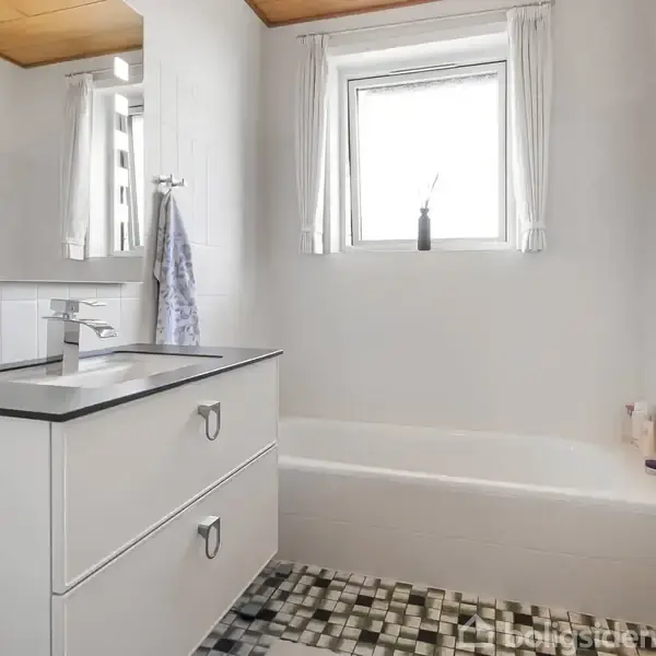 Badeværelse med hvidt skabsmodul med vask, spejl på væggen og badekar ved siden af et vindue. Flisebelagt gulv i sort-hvidt mønster. En radiator ses ved siden af badekarret.