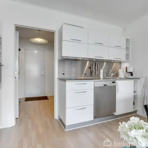 Køkken med hvide skabe og indbygget opvaskemaskine og ovn. Et rustfrit stållook dominerer bordpladen og bagvæggen. I baggrunden ses en åben dør til gangen. En buket hvide blomster står i forgrunden.