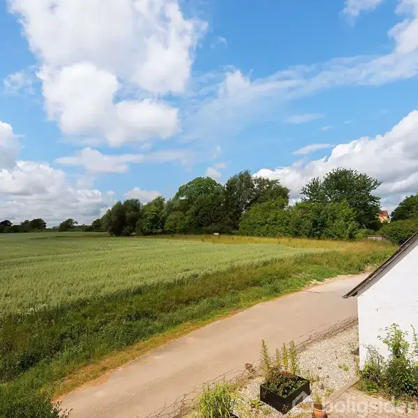 En hvid bygning står ved siden af en grusvej. Omkring bygningen er der grønne marker og træer under en blå himmel med spredte skyer.