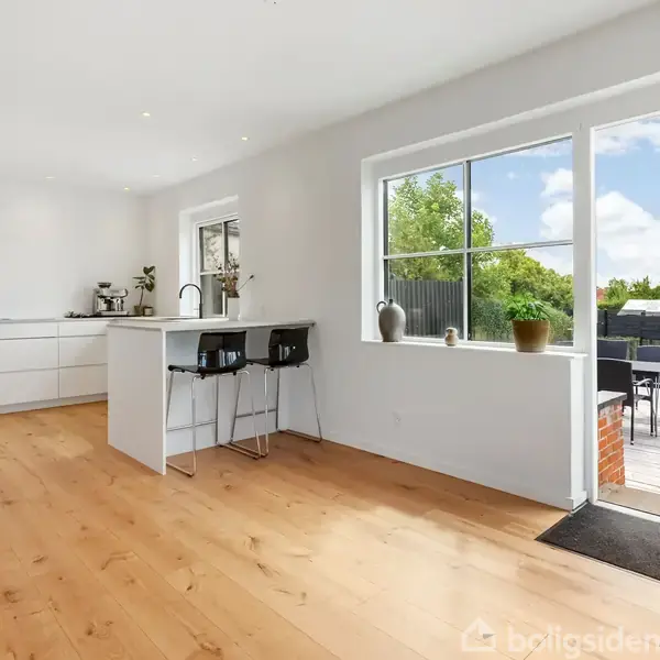 Moderne køkken med hvidt interiør og barstole ved en ø; store vinduer med udsigt til terrasse og grønne træer udenfor. Scenen er rolig og lys.