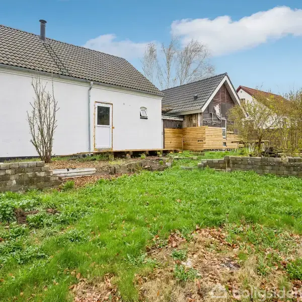 Et hvidt hus med skråt tag står i en græsbevokset have. Huset har en træterrasse ved indgangen, omgivet af buske og træer. En murstenstrappe leder op til terrassen.