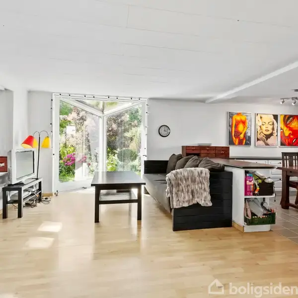 Stue med trægulv og store vinduer, der åbner ud til en have. Sofaer er placeret ved et sofabord. Spisebord og stole står nær farverige vægmalerier og en åben terrassedør.