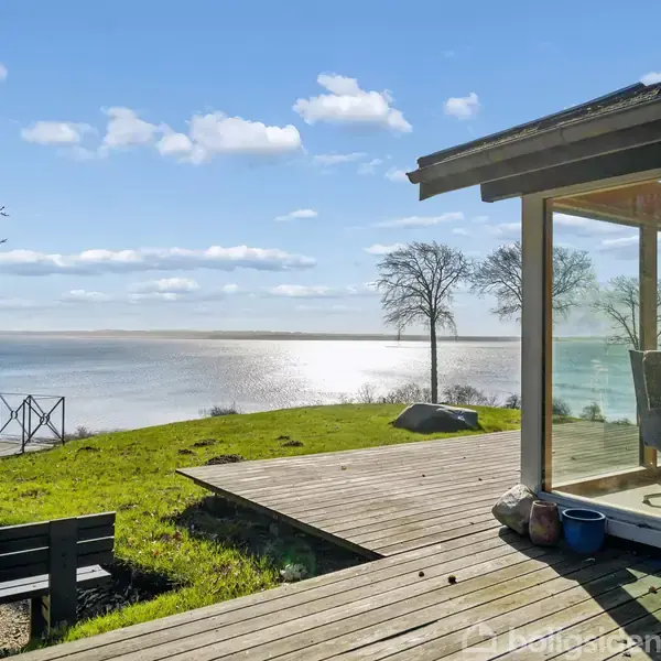 Et hus med glasvægge står ud mod en havudsigt. Sollys reflekteres på vandet. Der er en træterrasse, græsplæne, træer og en bænk i nærheden.