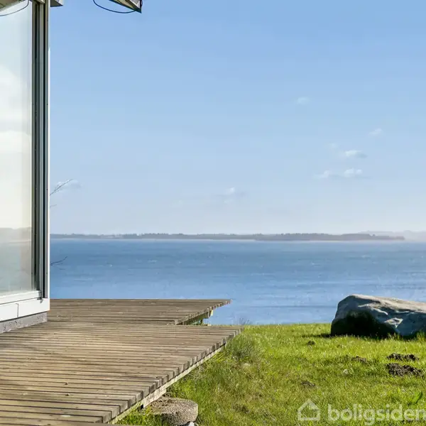 Glasfacade reflekterer sollys, stående ved en træterrasse. Udsigten strækker sig over en rolig blå fjord under en klar himmel, omgivet af en grøn græsplæne med en stor sten på.