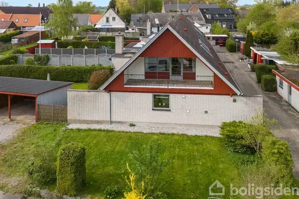 Et rødt og hvidt hus med et trekantet gavl, vinduer og altan, står i en forstad med hække og nabohuse, omgivet af en velholdt have og garage.