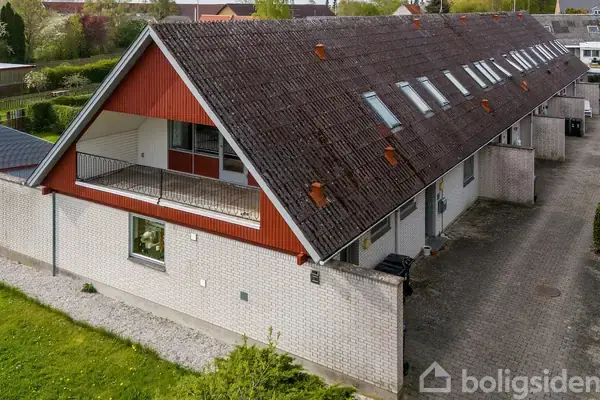 Rækkehus med skråt tag og røde detaljer, hvor rækkehusene ligger langs en flisebelagt sti. Græsplæner og træer omgiver huset i en rolig, beboelig område.