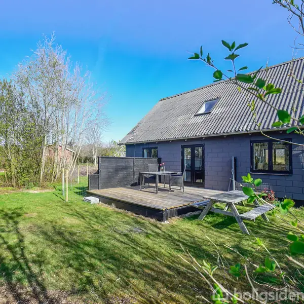 Et gråt træhus står i solskin med en træterrasse foran, omgivet af en grøn have med træer og græs. Der er ingen tekst bortset fra "Boligsiden".