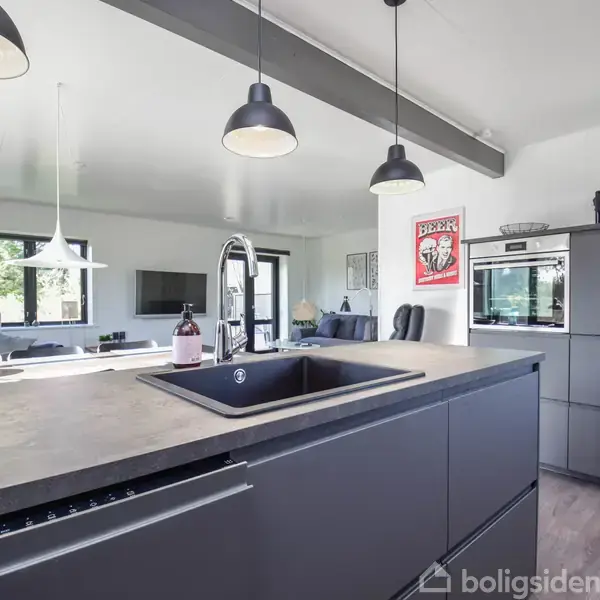 Køkkenø med mørk vask står i moderne køkken; hængelamper ovenfor, sort ovn og mikrobølgeovn indbygget i skab, lys træstruktur, plakat med teksten "Beer Helps Ugly People..." på væg.