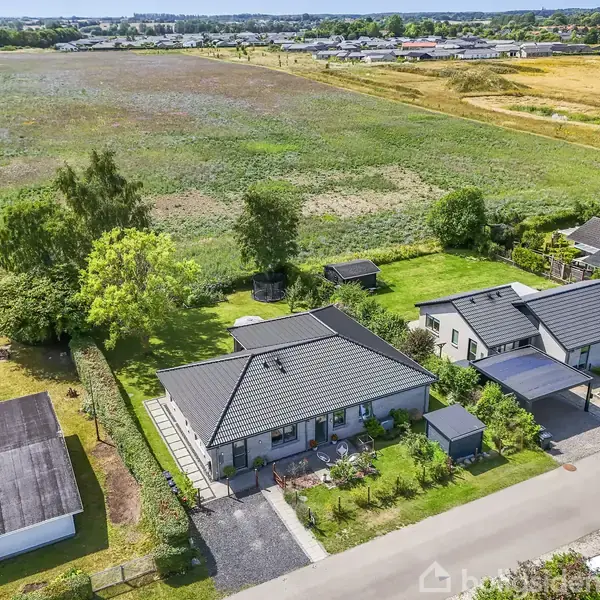 En moderne villa med gråt tag og velplejet have ligger fredeligt langs en stille vej. Bagved strækker en stor mark sig ud, omkranset af flere træer, under en klar himmel.