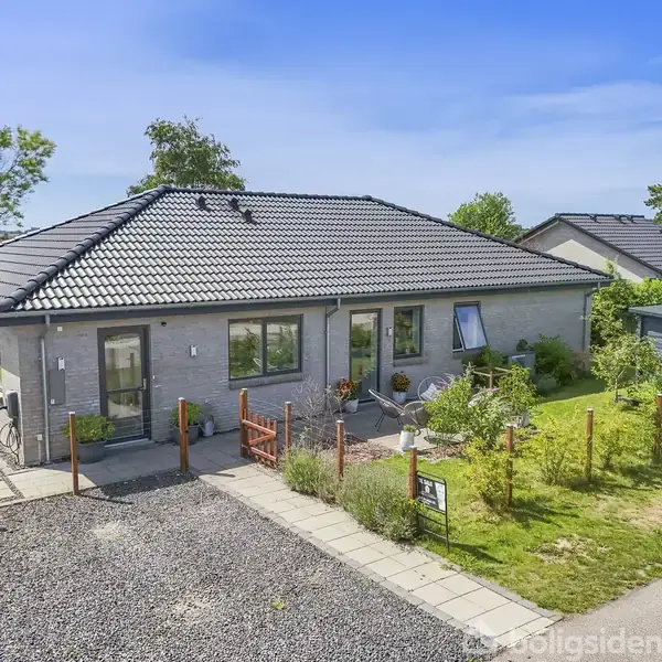 En moderne villa med sort tegltag står roligt. Foran er en velholdt have med blomster og en flisebelagt gangsti. Omkring er der grønne træer og en grusbelagt indkørsel.