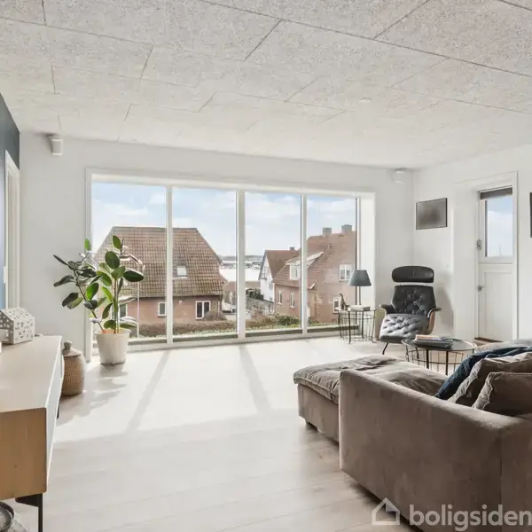En lys stue med sofa og lænestol, store vinduer mod en udsigt over tage. Der er potteplanter og et træmøbel med dekorative elementer langs den venstre væg.