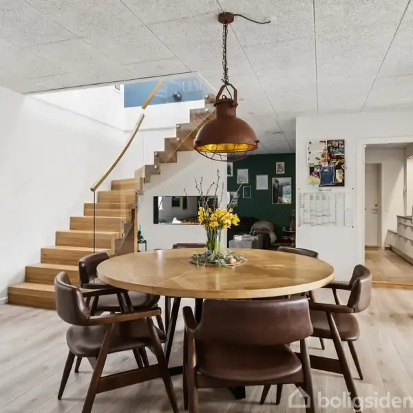 Rundt spisebord med brune stole står i et lyst rum. Ovenover hænger en rustik lampe. Ved siden af ses en trappe, og en stor urpyntet væg. Der hænger kunst på væggen.