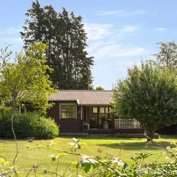 Et lille træhus med en veranda står i en frodig have. Træer og buske omgiver huset, og en klar blå himmel ses i baggrunden, skaber en fredfyldt atmosfære.