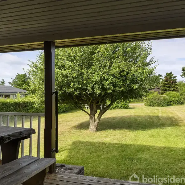 Træbord på en overdækket terrasse står stille. I baggrunden ses en græsplæne med et stort træ. Der er også andre huse og flere træer i horisonten.