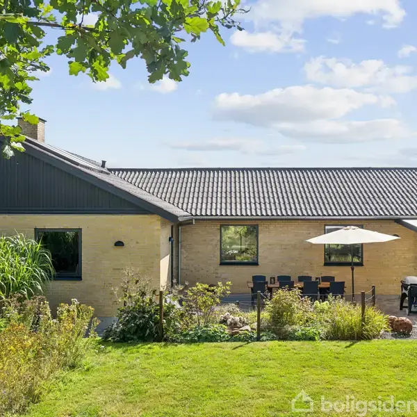 Hus med gul murstensvægge og sort tag står i en have. En terrasse med spisebord og parasol ligger foran bygningen, omgivet af grønne planter og en veltrimmet græsplæne under en blå himmel.
