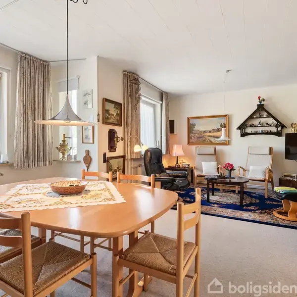 Spisebord med seks stole står i et lyst rum. Der er en loftslampe over bordet. Stuen har bogreoler, en sofa, lænestole og tæpper. Store vinduer lader lys ind.