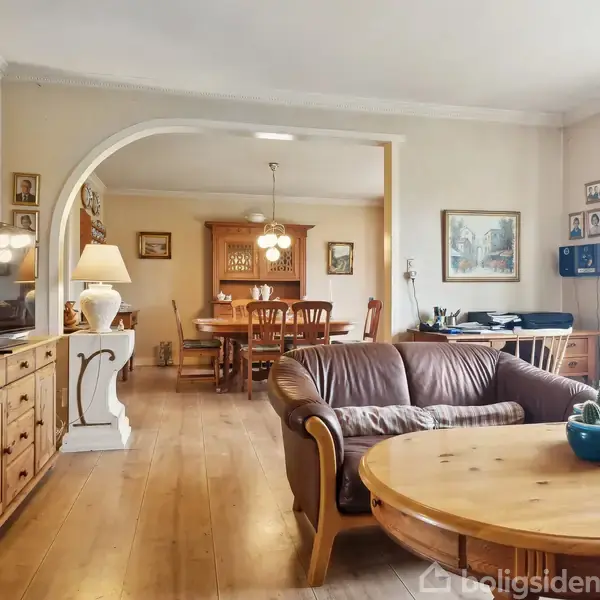 Stue med brunt lædersofa-sæt foran et rundt sofabord; en fladskærms-TV står på et træskab. I baggrunden ses en lys spisestue med træmøbler under en lysekrone.
