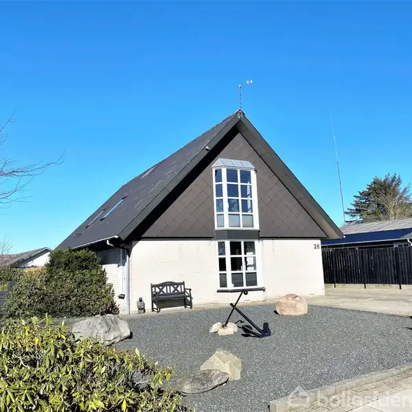 En A-formet hus med store vinduer og sort tag står i en grusbelagt forhave med dekorative sten. Skiltet "Til Salg" er placeret i indkørslen. Himmel og buske omkranser.