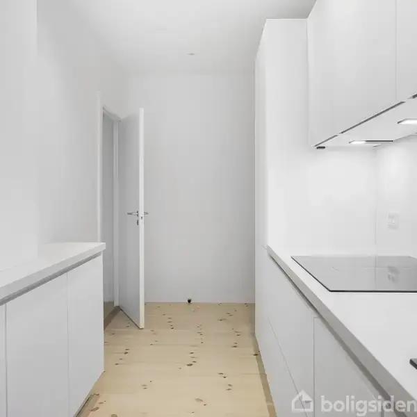 Et lyst køkken med hvide skabe og fliser, hvor der er installeret en indbygget kogeplade. En åben dør fører til en anden del af huset. Gulvet er lyst trægulv.