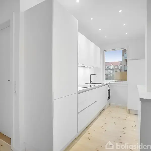 Et moderne, hvidt køkken med skabslåger, vask og vaskemaskine i brug. I baggrunden ses et vindue, der lukker dagslys ind, med udsigt til nabobygninger.