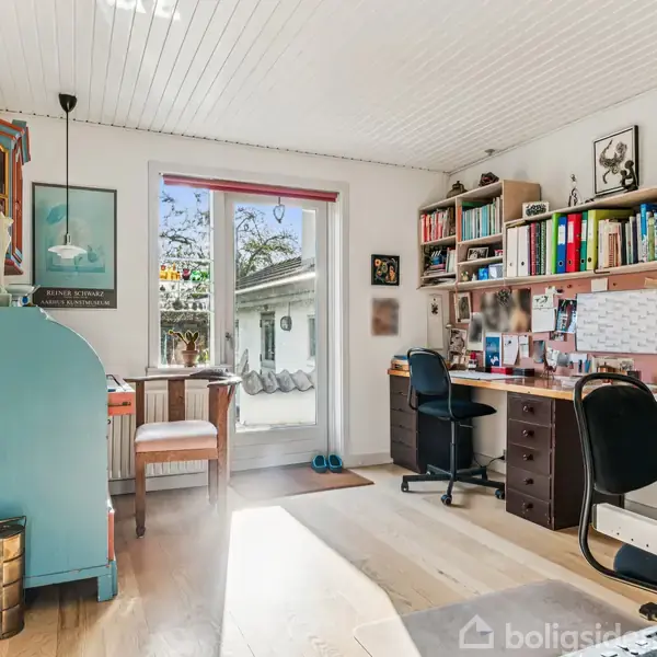 Et rummeligt arbejdsrum med skrivebord og hylder fyldt med bøger og papirer, to skrivebordsstole. Et vindue og dør fører ud til en udendørs gård med træer. Ingen tekst.