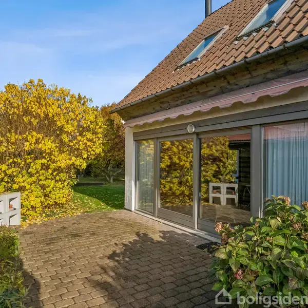 Huset har en stor glasdør, der åbner ud mod en flisebelagt terrasse. En markise hænger over døren. Træer i efterårsfarver omgiver området.