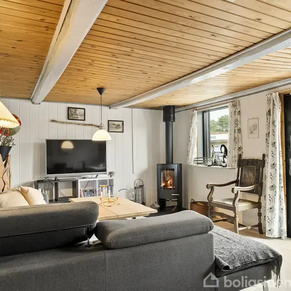 En stue med en grå sofaarrangement ved et sofabord, en tændt brændeovn ved siden af vinduer med gardiner. Fjernsynet står mod væggen, dekoreret med billeder og en lampe.