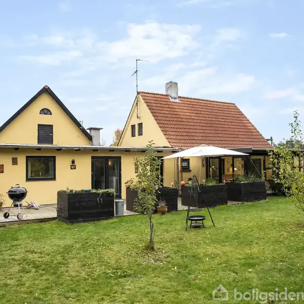 Et gult hus med rødt tag står omgivet af en grøn have. En grill og parasol placeret på terrassen danner en hyggelig udendørs opholdsplads.