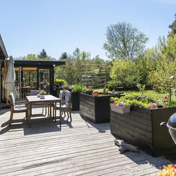 Terrasse med træbord og stole, omgivet af frodige planter og træer. En grill står ved siden af plantekasser. Sollys oplyser området, der skaber et indbydende udendørsmiljø.