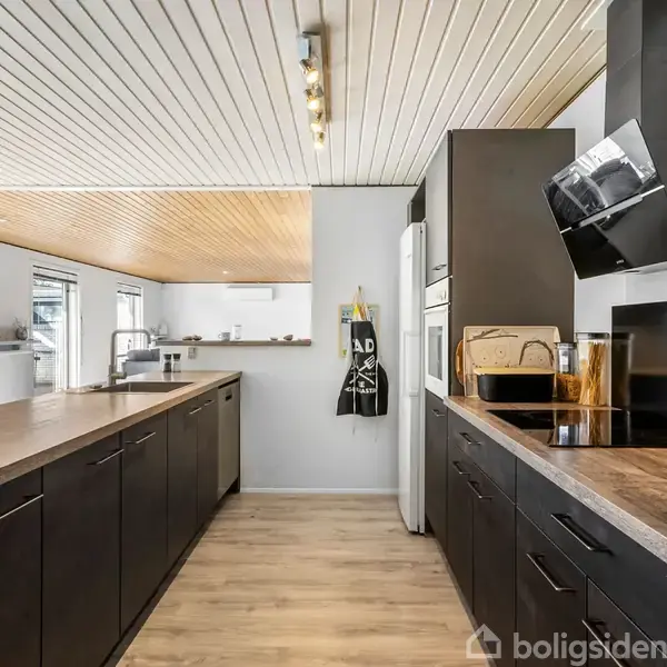 Køkken med sorte skabe og træbordplader, placeret i et rum med trægulv og hvidt loft med spotlys. Foran er der åbne vinduer og moderne apparater. En forklæde hænger på væggen.