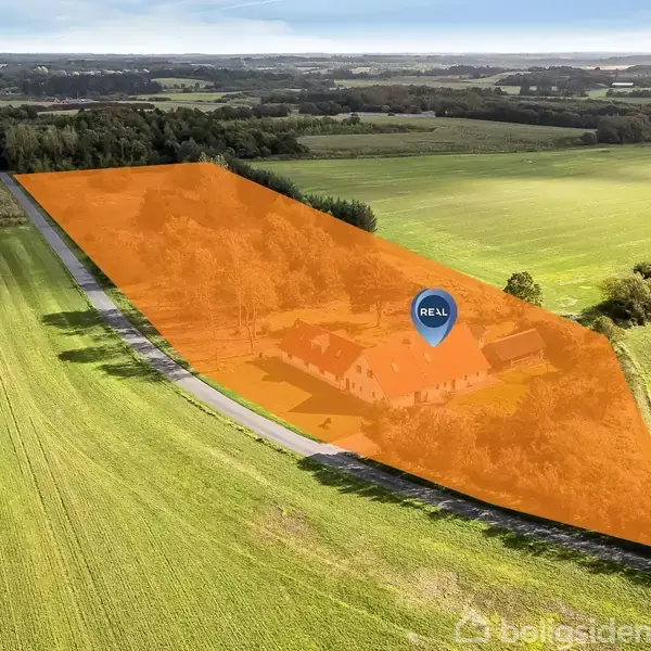 En orange markering viser en ejendom med en bygning i midten, omgivet af åbne grønne marker og skov. En blå markør med teksten "REAL" vises over bygningen.