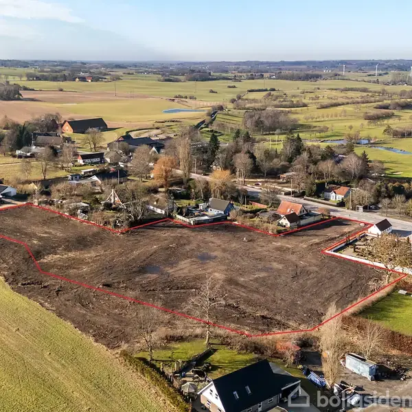 En stor byggegrund markeret med rød linje ligger i en landlig landsby, omgivet af huse, veje og åbne marker under en klar himmel.