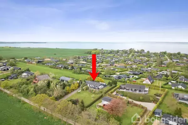 En rød pil peger på et hus i en landlig bebyggelse omgivet af grønne marker og træer nær en sø i baggrunden.