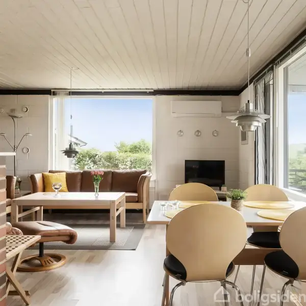 Stue med brunt sofaarrangement om et kaffebord, store vinduer, og spiseplads med lysbord. Kamin mod væggen og moderne belysning over bordet skaber en hyggelig atmosfære.