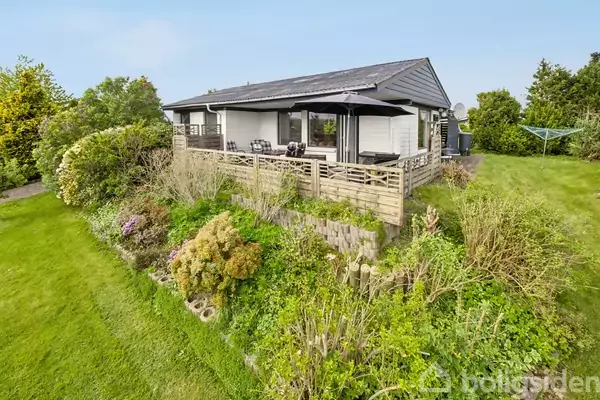 En moderne bolig har en terrasse med møbler og en parasol, omgivet af frodig have med græsplæne og buske i en rolig, grøn beliggenhed.