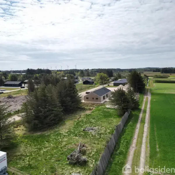 Et hus står stille omgivet af træer og marker. Langs en grusvej ses flere huse spredt ud over et grønt landskab med vindmøller i horisonten under en overskyet himmel.