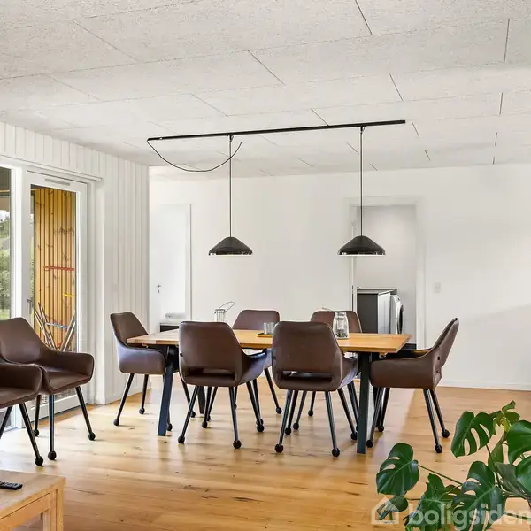 Brune læderstole står omkring et træspisebord, oplyst af moderne, sorte pendellamper. Rummet er lyst med hvidmalede vægge og vinduer, med grønne planter tilføjer naturlig kontrast.