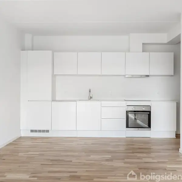 Et køkken i moderne stil har hvide skabe og en ovn, placeret mod en hvid væg, med trægulve som en del af et åbent rum.