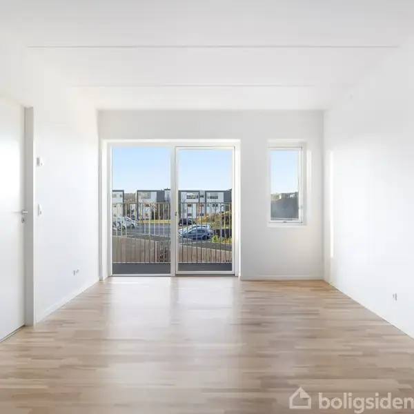 Et tomt rum med lyse trægulve og hvide vægge. En glasdør åbner til en balkon med udsigt over en bygningskompleks. Sollys strømmer ind gennem vinduerne.