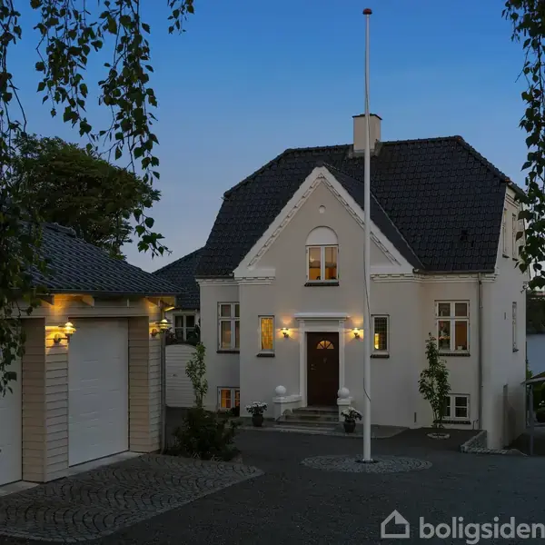 En hvid villa med sort tag står oplyst ved skumring. Til venstre ses en carport. I baggrunden skimtes en sø omgivet af træer.