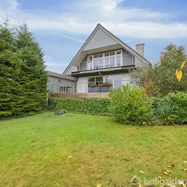 Et moderne hus med balkon står stille omgivet af en velplejet have med buske og træer. Skyfri himmel i baggrunden.