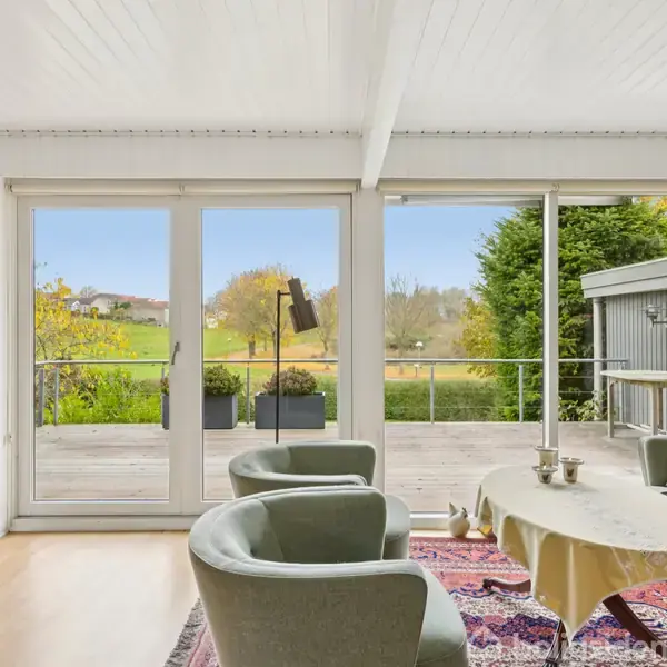 Stue med to grønne stole og et bord med dug, står foran store glasdøre, der åbner ud til en terrasse med udsigt over en grøn have og landskab.