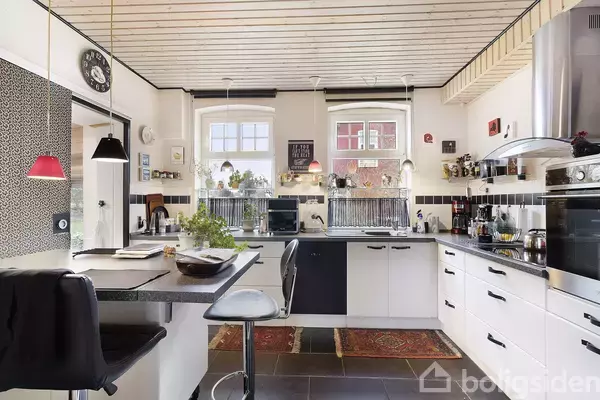 Et moderne køkken med hvide skabe og sort bordplade. Barstole står ved en morgenmadsbar. Vinduer slipper lys ind, og der er grønne planter i vindueskarmen. En klokke hænger på væggen.