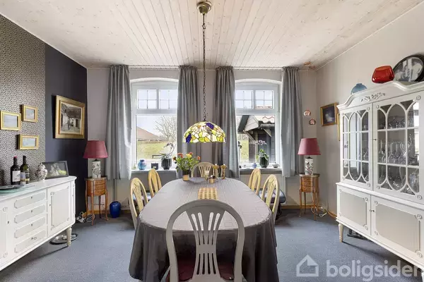 Spisebord med dug og blomsterarrangement under en farvet lampeskærm i en spisestue med lyse skråvægge, vinduer med grå gardiner og antikke møbler samt pynteting på skabe.